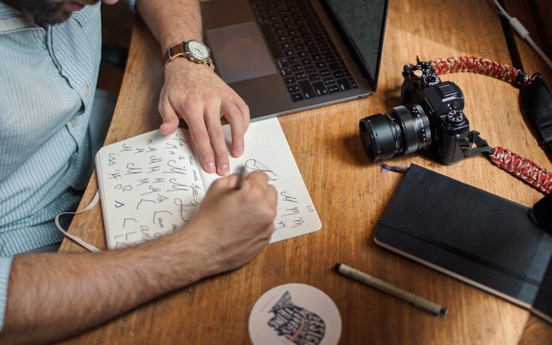 Person schreibt auf Papier mit Laptop, Kamera und Notizbuch auf dem Tisch.
